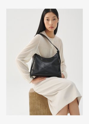 Young woman with long black hair sits on wooden block wearing cream outfit and holds black leather shoulder bag across chest.