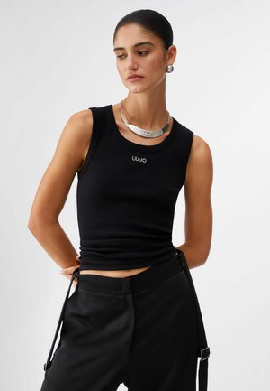 Frau trägt ein schwarzes ärmelloses Oberteil mit dem "LIU JO"-Logo, schwarze Hosen mit seitlichen Riemen, silberne Halskette und Creolen, schaut seitlich.