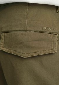 Pantaloni in tessuto verde oliva con una tasca posteriore con patta. Logo "GANT" ricamato sopra la tasca in una tonalità discreta. Texture liscia.