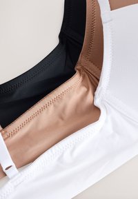 Trzy bezszwowe staniki w czerni, beżu i bieli ułożone na sobie, aby podkreślić szwy i gładką strukturę tkaniny.