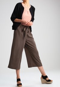 Kvinna iförd en svart öppen cardigan, en rosa topp med spetskant, lösa bruna cropped byxor och svarta kilskor, poserande mot en ljus bakgrund.