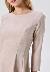 Robe beige en tissu doux et texturé avec un motif en chevrons, dotée d'un col rond et de manches trois-quarts.