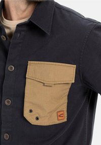 Mørkeblå skjorte med en tan farvet fløjl brystlomme, der har en klap, to øjer og et logo-patch. Knappedyrknap foran med brune knapper.