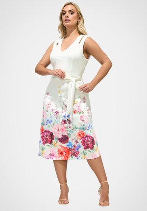 Kobieta stoi, ubrana w białą, bezrękawnikową sukienkę z kolorowym kwiatowym wzorem na spódnicy, związaną paskiem, oraz beżowe szpilki z paskiem wokół kostki na białym tle.