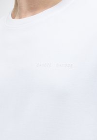 Κοντινό πλάνο ενός ατόμου που φοράει απλό λευκό T-shirt με διακριτικό ανάγλυφο κείμενο "SAMSØE SAMSØE" στο στήθος.