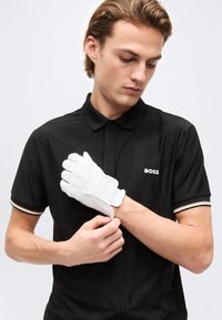 Νέος άνδρας με μαύρο πόλο BOSS ρυθμίζει ένα λευκό γάντι γκολφ στο αριστερό του χέρι σε απλό φόντο.