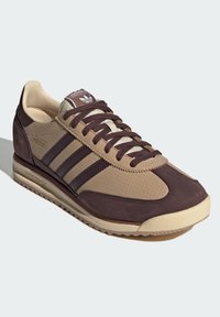 Chaussure de sport marron et beige avec textile perforé et suède, dotée de trois bandes, d'un laçage à l'avant et d'une semelle en caoutchouc texturée.