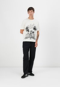 T-shirt en coton blanc avec un motif graphique noir représentant des palmiers et une vue de la ville, associé à un pantalon large noir et des baskets noires.