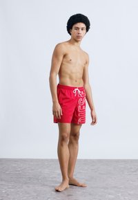 Punased ujumispüksid, mis on valmistatud kerge materjali ning millel on valge "DIESEL" logo-disain, elastne vöökoht ja paelaga kinnitamine.