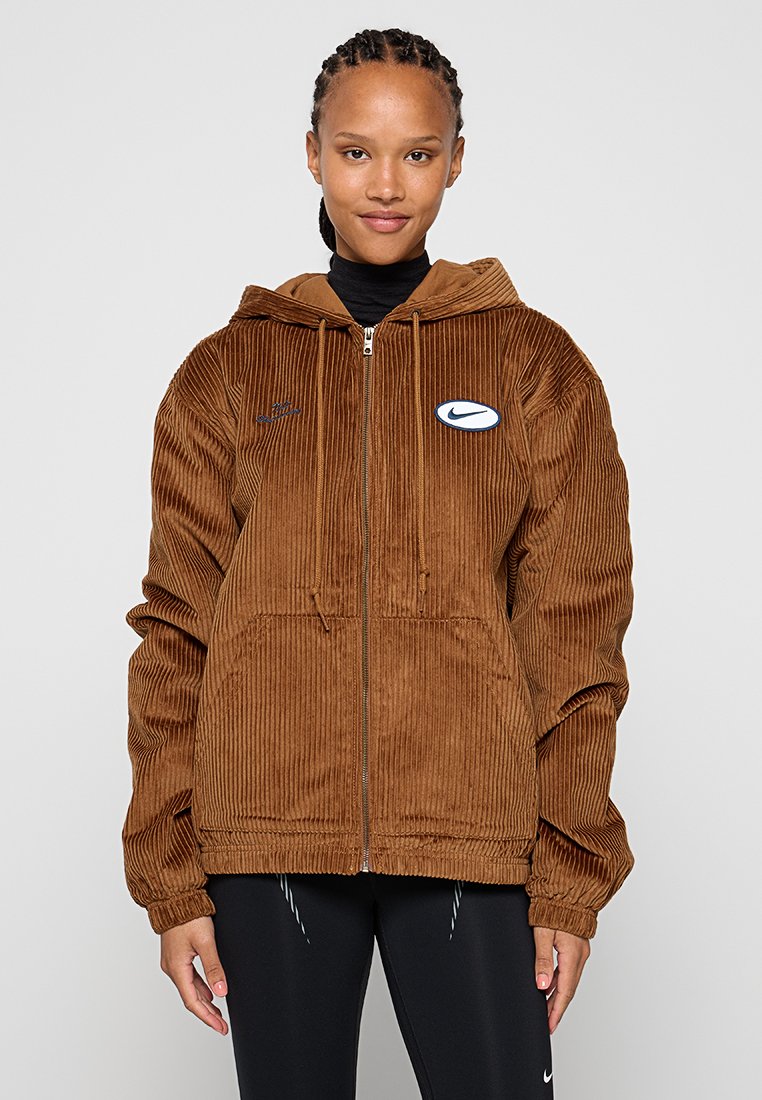 Chaqueta con capucha de pana marrón con cierre de cremallera, bolsillos frontales y un prominente logo azul de Nike en el área del pecho izquierdo. Tejido texturizado.