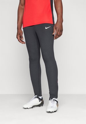Leggings atletici grigi a costine con logo Nike, abbinati a una maglietta a maniche corte rossa e nera e scarpe da calcio bianche.