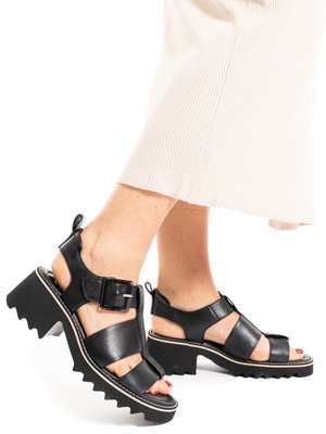 Füße, die schwarze Sandalen mit klobigen Absätzen, dicken Riemen und Schnallen tragen, kombiniert mit hellen, weit geschnittenen Hosen.