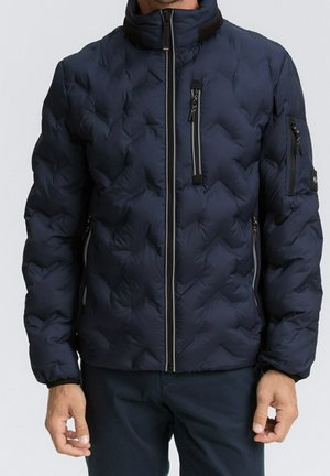 Mann trägt eine marineblaue gesteppte Jacke mit Reißverschlusstaschen vorne und an den Ärmeln, steht mit entspannt an den Seiten hängenden Händen.