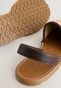 Sandale en cuir marron avec une bride épaisse, une semelle cousue et une semelle extérieure en caoutchouc texturée avec motif de bande de roulement géométrique sur fond blanc.