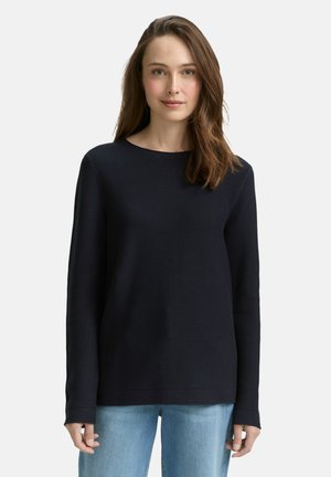 Mujer con cabello largo y castaño, vestida con un suéter azul marino de manga larga y acanalado, y unos vaqueros azul claro, de pie frente a un fondo claro y liso.