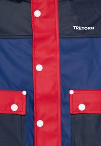 Wasserdichte Jacke in einem navy-blauen und roten Farbschema, mit weißen Druckknöpfen und zwei Fronttaschen. Hergestellt aus glattem, strapazierfähigem Material.