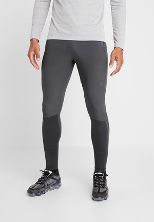 Mężczyzna w ciemnoszarych obcisłych spodniach sportowych i czarnych butach sportowych Nike, stoi z nogami lekko rozstawionymi na jednolitym tle.