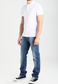 Hombre vestido con una camiseta polo blanca de manga corta, jeans azules y zapatillas en blanco y negro, de pie frente a un fondo claro y liso.