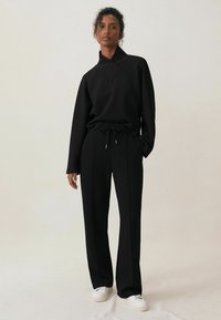 Zwart loungewear set met een langemouwtop met hoge kraag en broek met trekkoord. Soepele textuur en een ontspannen pasvorm, gecombineerd met witte sneakers.