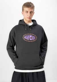 Jonge man in een zwarte Rip Curl hoodie met een paars en roze logo, handen in de zakken, staand tegen een eenvoudige witte achtergrond.
