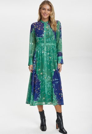 Langes, knielanges Hemdkleid in Grün und Blau mit floralen und geometrischen Mustern. Es hat lange Ärmel und einen schwingenden Rock und wird mit schwarzen Knöchelboots kombiniert.