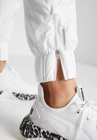 Bílé teplákové manžety na zip, s elastickým lemem a reflexním detailem. Kombinováno s bílými teniskami s černou podrážkou se vzorem leopardího tisku.