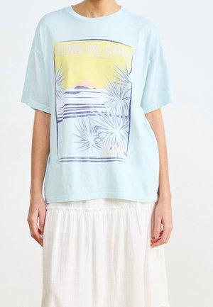 Osoba nosząca jasnoniebieski oversize t-shirt z napisem "COASTAL GOLD" i grafiką przedstawiającą nadmorski pejzaż, zestawiony z białą zwiewną spódnicą.