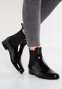 Bottes de cheville en caoutchouc noir avec panneaux élastiques latéraux, bout rond et petit talon carré, présentées à côté de jeans skinny gris.