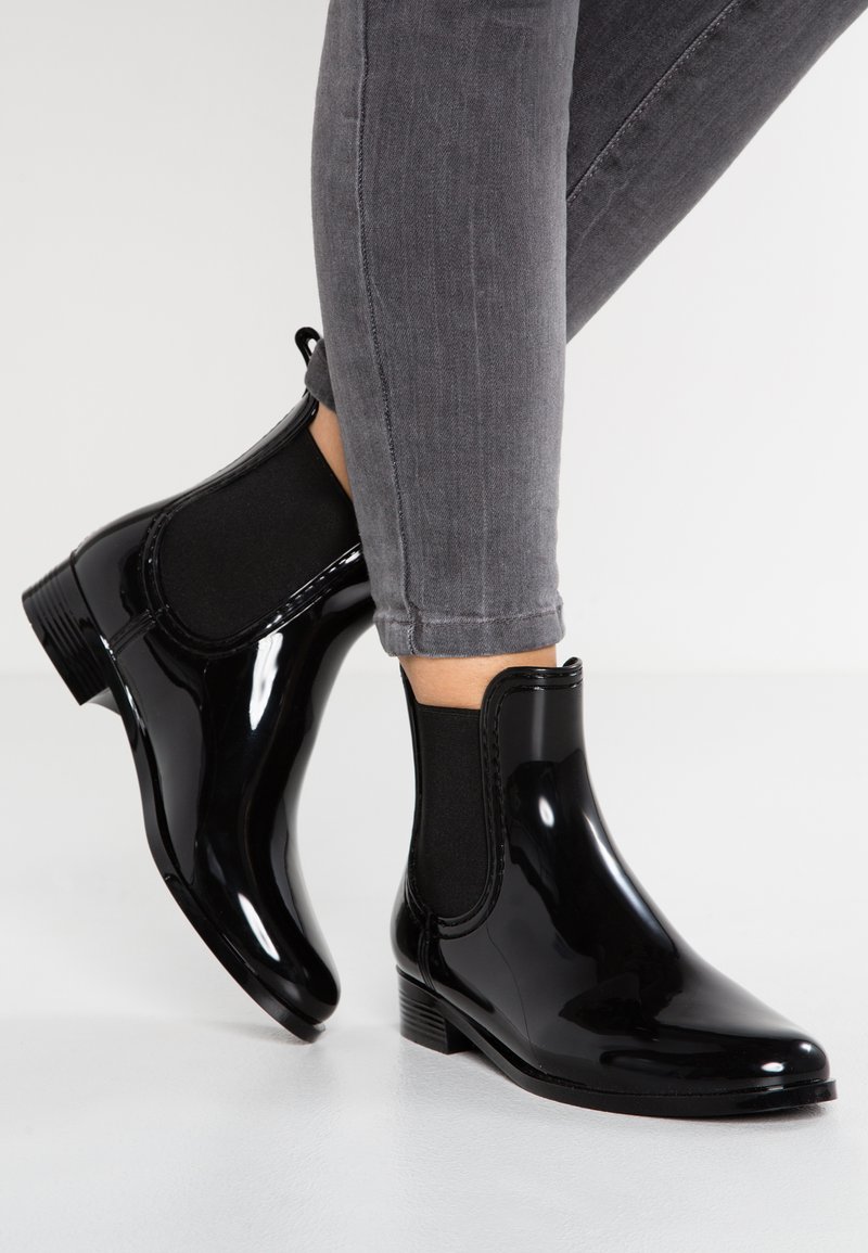 Bottes de cheville en caoutchouc noir avec panneaux élastiques latéraux, bout rond et petit talon carré, présentées à côté de jeans skinny gris.