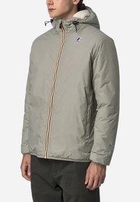 Giacca isolante grigio chiaro con zip frontale, cappuccio con cordoncino e rivestimento interno morbido. Presenta zip bicolore e patch con logo.