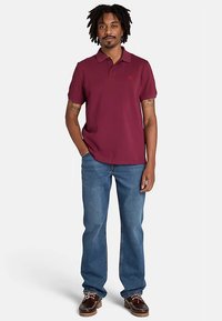 Tummanpunainen poolopaita, lyhyet hihat, pienellä logolla rinnassa. Yhdistetty sinisiin farkkuihin ja ruskeisiin kenkiin. Malli seisoo kädet taskuissa.