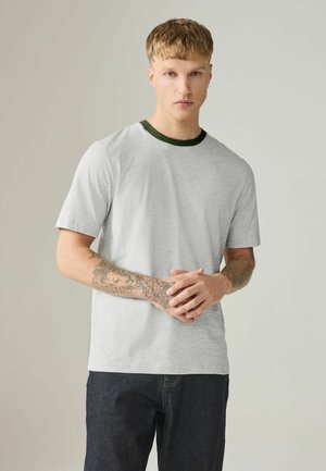 Ung mand med kort blondt hår iført en lysegrå T-shirt med mørk krave og mørke jeans, hænder foldet, der viser tatoveringer på underarmene.