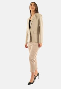 Blazer beige con solapa normal y dos bolsillos frontales, combinado con pantalones beige claros y tacones altos negros y brillantes, mostrado sobre un fondo blanco.