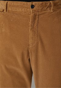 Detailopname van de voorkant van bruine corduroy broek met zichtbare riemlussen, knoopsluiting en gestructureerde verticale ribbels.