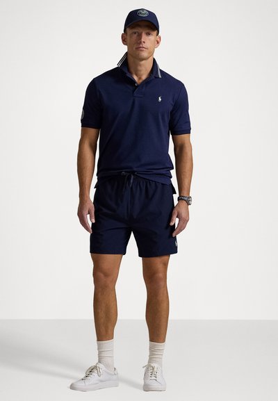 Polo shirt bleumarin cu guler în dungi, asortat cu pantaloni scurți bleumarin. Adidași albi și șosete completează ținuta, evidențiind un aspect sport.