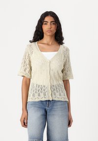 Knit cardigan in cream with a V-neck, short sleeves, and a floral lace pattern. Features buttons down the front and a slightly sheer texture.