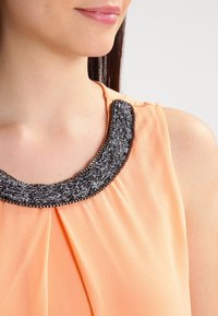 Ärmlös topp i persikofärgad tyg med en svart vävd detalj vid halsringningen. Tyget är mjukt med veck vid bröstområdet, vilket skapar mjuka veck.