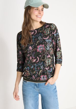 Lächelnde Frau mit grüner Mütze, schwarzem Langarmshirt mit Blumenmuster und hellblauer Jeans, stehend vor einem schlichten weißen Hintergrund.