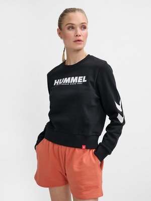 Mujer con coleta rubia vistiendo sudadera negra de Hummel y pantalones cortos coral, con las manos en los bolsillos, mirando hacia su izquierda sobre fondo blanco.