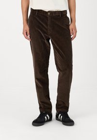 Brune kordfløyelsbukser med vertikale ribber, rett ben design, knappelukking og sidelommer, sammen med svarte joggesko med hvite striper.
