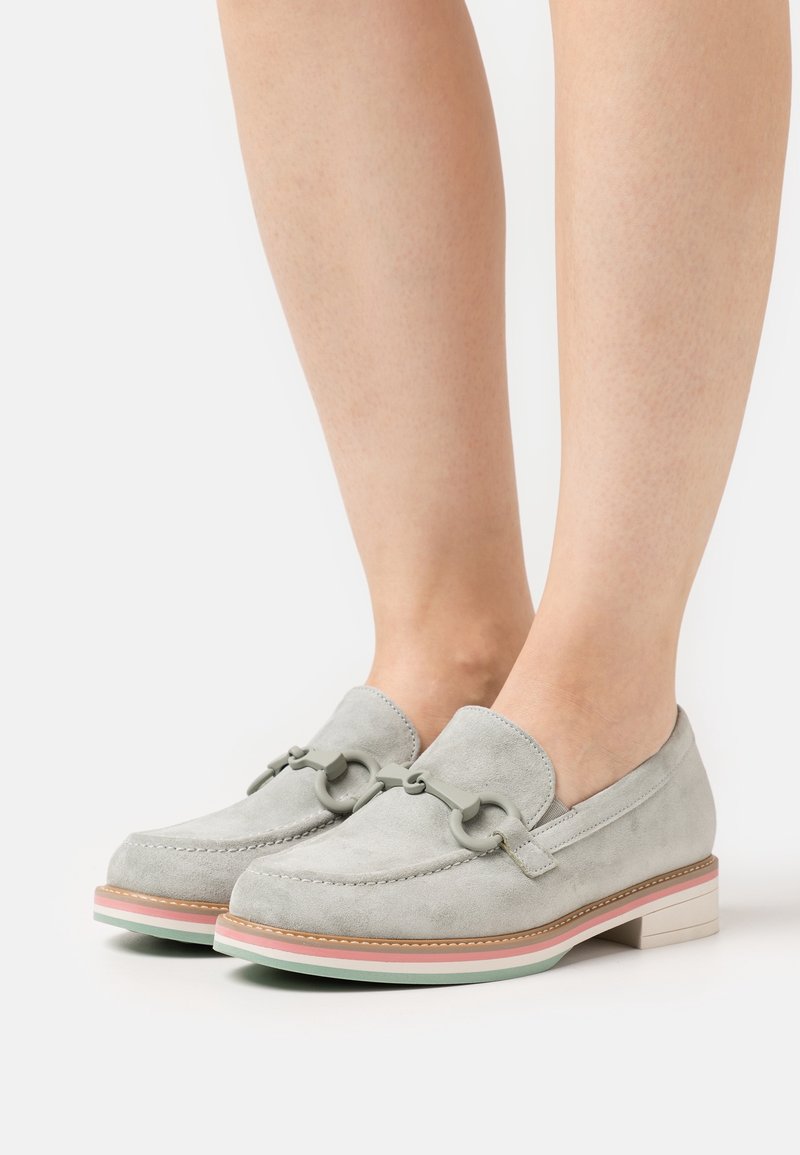 Mocassins en daim gris clair avec semelles rayées pastel et boucles décoratives grises, portés aux pieds sur un fond blanc.