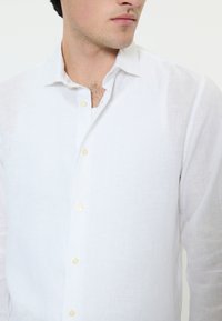 Moški v beli laneni srajci z dolgimi rokavi in gumbi, ki gleda v stran, z vidnimi tremi gumbi in rahlo odprtim ovratnikom.