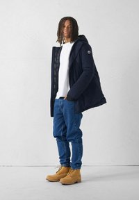 Giovane con dreadlocks che indossa una giacca a vento blu navy, una camicia bianca, jeans blu e stivali beige con lacci, in piedi contro un muro bianco.