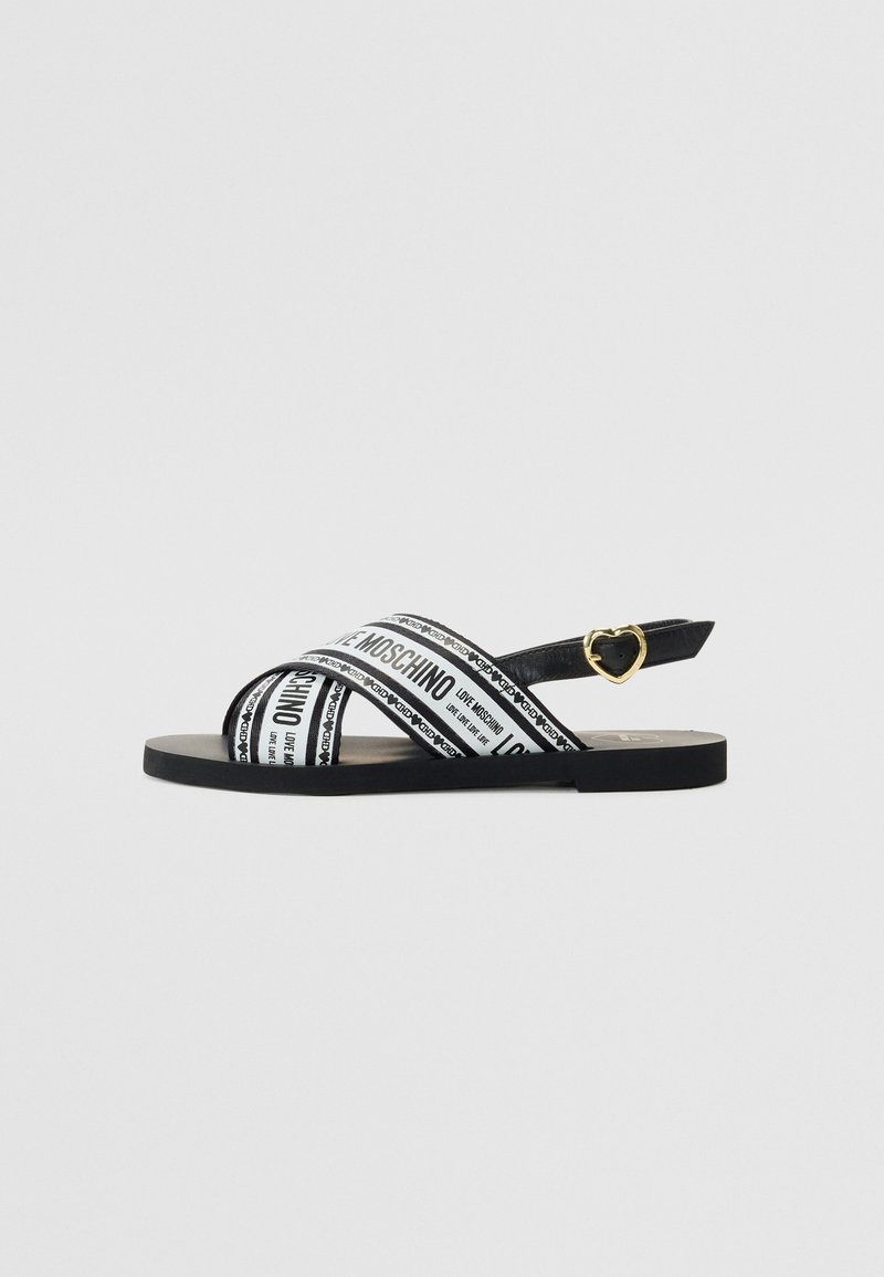 Sandal preto com tiras cruzadas apresentando texto branco. Sola plana, detalhe em forma de coração em hardware dourado na tira do tornozelo.