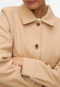 Femme portant un manteau en laine beige avec de grands boutons, les bras croisés, et une boucle d'oreille pendante visible d'un côté.