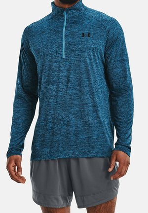 Homme portant un haut de sport bleu à zip quart et manches longues et un short de sport gris, debout les bras détendus le long du corps.