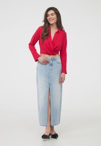Camicia rossa corta con bottoni e maniche lunghe, abbinata a una maxi gonna in denim azzurro chiaro con spacco frontale e orlo sfilacciato. Scarpe nere con dettaglio a fiocco.