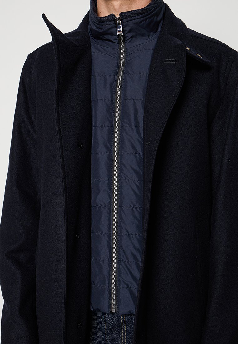 Cappotto di lana nera sovrapposto a una giacca a zip trapuntata blu navy. Presenta un colletto alto, due bottoni e un design elegante e minimalista.