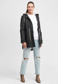 Zwarte waterdichte parka met capuchon, gevoerde kraag, rits en verstelbare details, gecombineerd met lichtblauwe gescheurde jeans en enkellaarsjes met slangenprint.