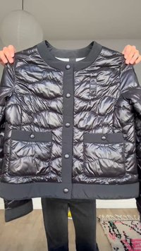 Ročaji držijo sijočo črno prešito jakno z zaponkami na pritisni gumb in dvema žepoma spredaj v svetli sobi z preprogo in papirnato lanterno.